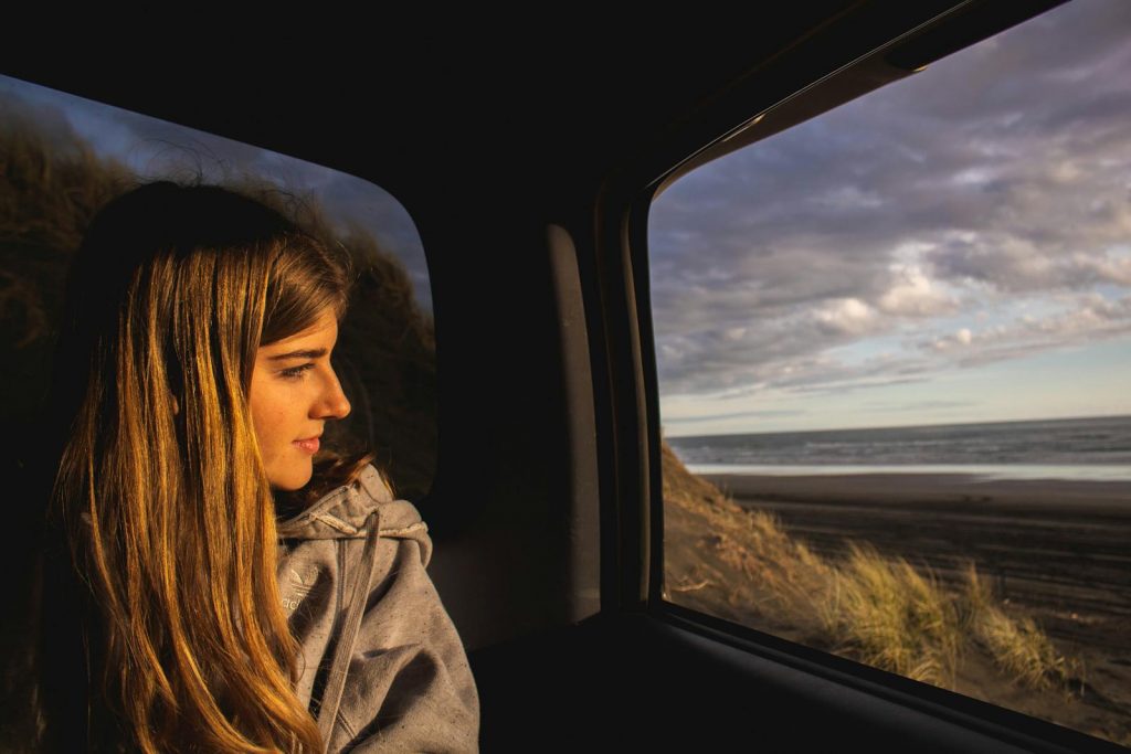 A young woman gazes out from a car window, appreciating a scenic beach view during daytime.